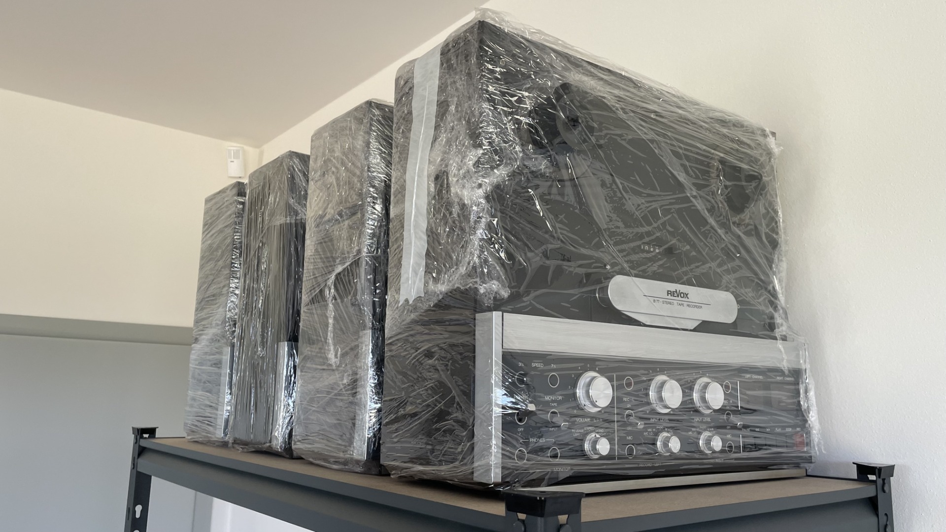 Zabalené audio zařízení v průhledné fólii na regálu, vidět ovládací panel a nápis Revox.